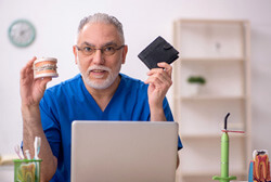an older male dentist holding up a model jaw and a wallet